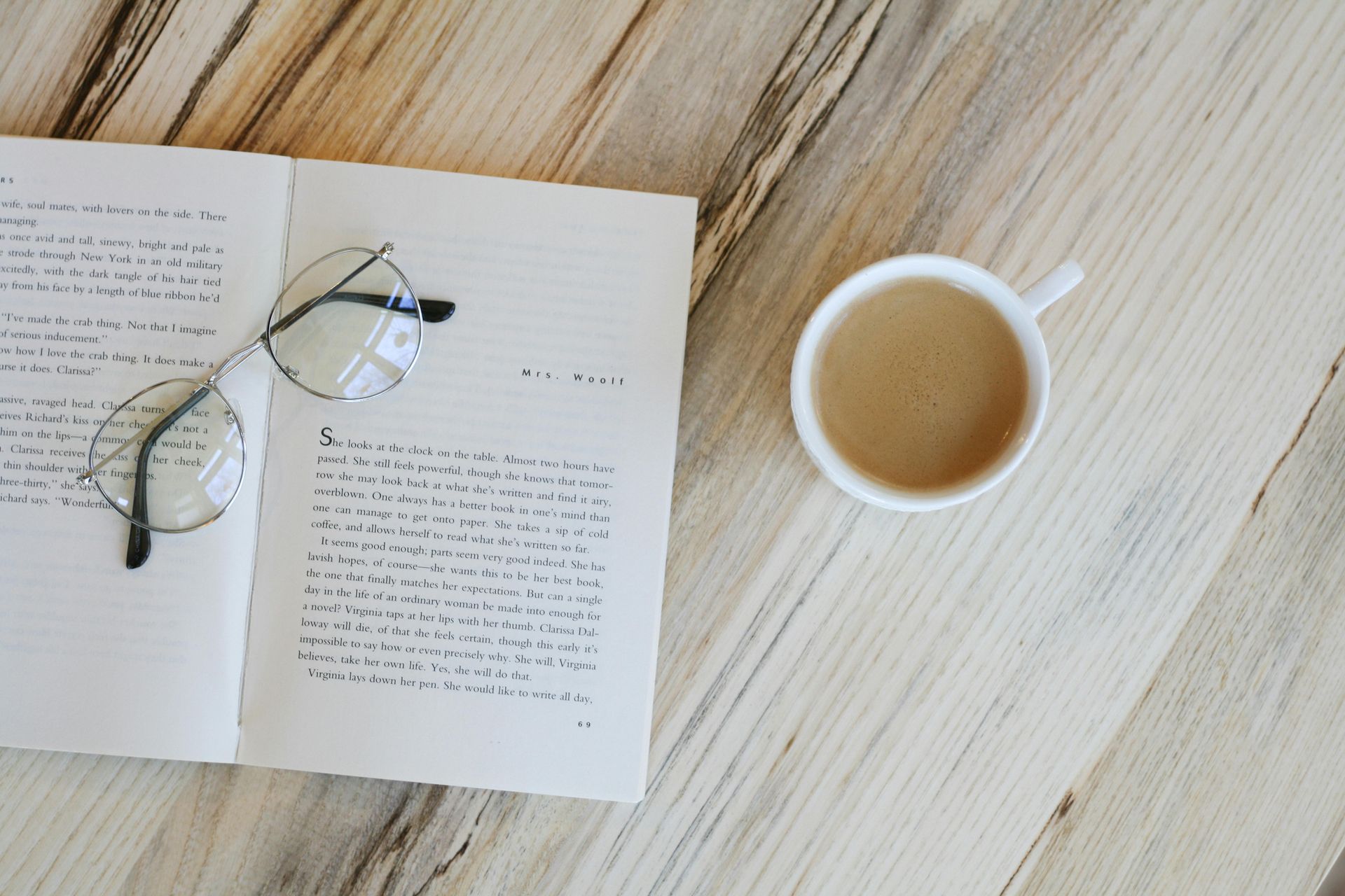 An open notebook with handwritten text and a pair of round glasses, beside a white mug of coffee on a wooden table.