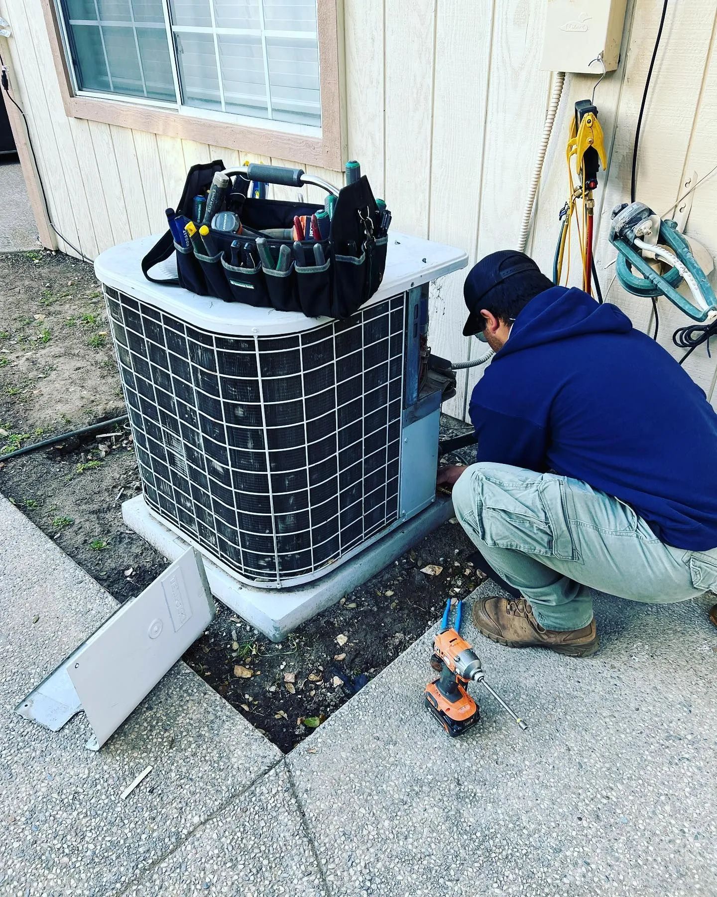 Technician kneels beside an outdoor air conditioner unit, using tools to service it by a house wall.