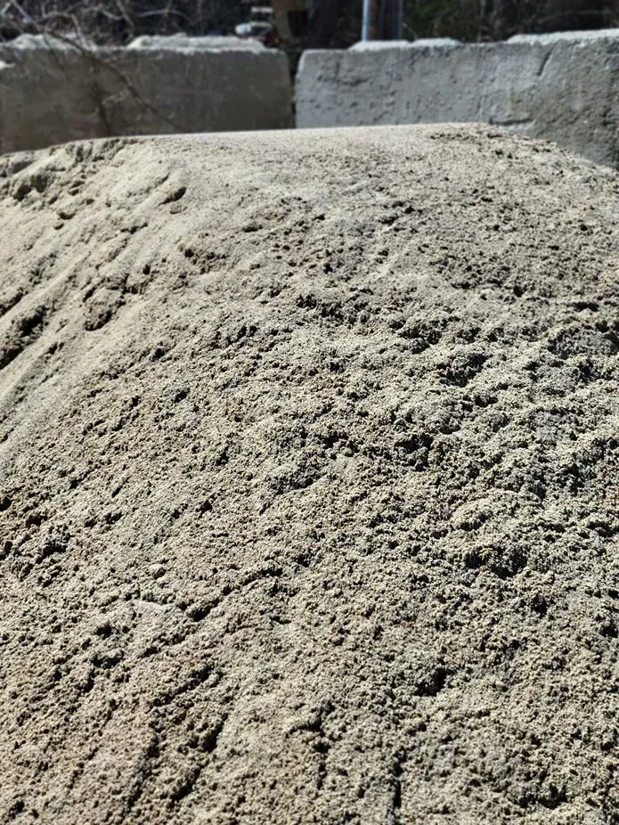 A large pile of coarse, light brown construction sand sits outdoors in front of grey concrete barriers.