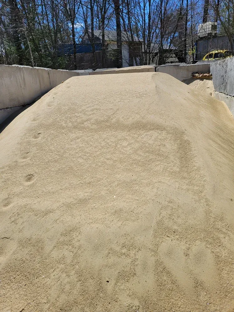A large, mounded pile of light-colored sand sits within a concrete storage enclosure surrounded by trees.