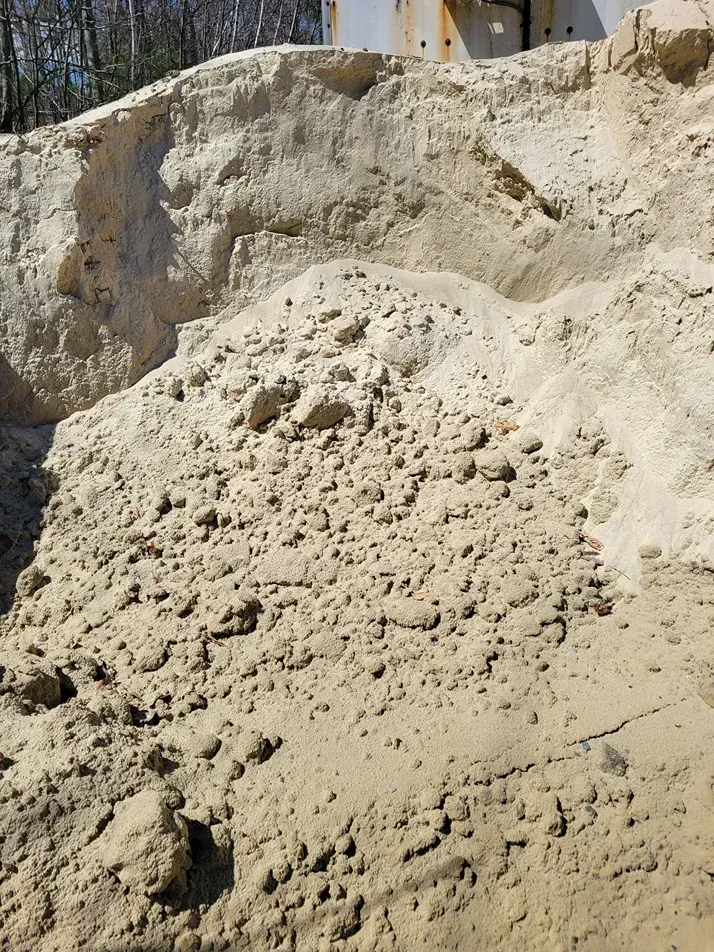 Pile of light tan sand or dirt outdoors, with rough mounds and a building edge in the background