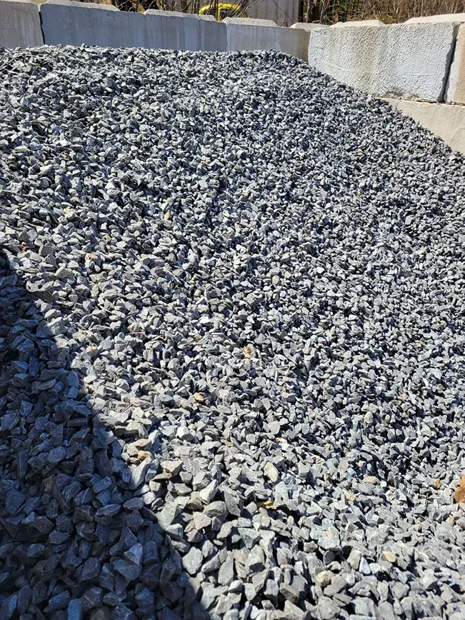 A large pile of uniform, dark gray crushed stone or gravel held within concrete barriers under bright sunlight.