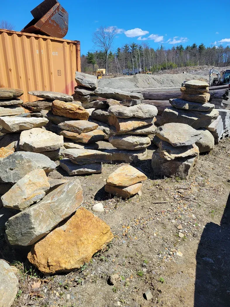 Stacked stones in a rocky outdoor yard beside a rust-colored shipping container under a blue sky