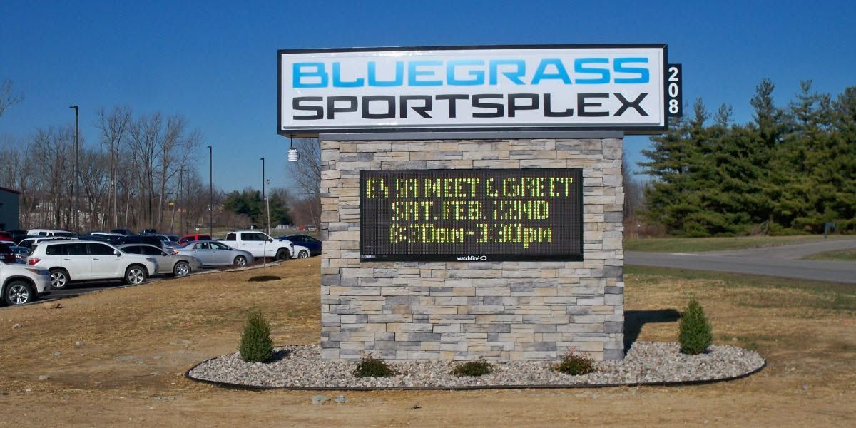 Bluegrass Sportsplex sign on a stone base beside a parking lot and trees under a clear sky. Bluegrass Sportsplex sign on a stone base beside a parking lot and trees under a clear sky.