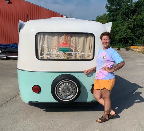 Woman posing beside a mint-and-white teardrop camper with a window curtain and spare wheel.