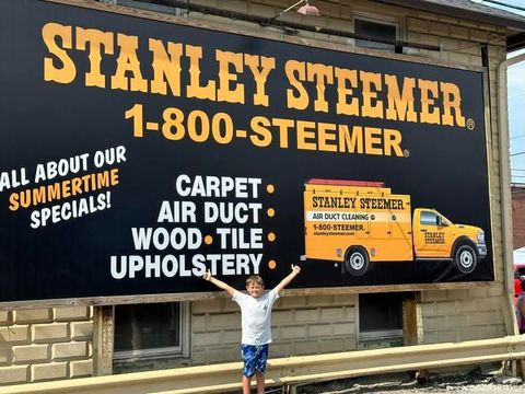 Child standing before a Stanley Steemer truck sign advertising carpet, air duct, wood, and tile upholstery cleaning.