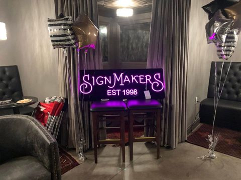 Neon pink “Signmakers EST 1996” sign in a room with two silver balloons and tables by curtains.