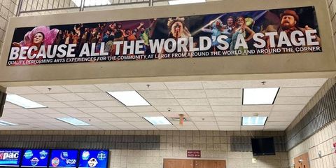 Banner in a hallway reads “Because all the world’s a stage” above ticket counters and ceiling lights.