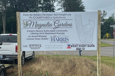 Outdoor sign for Magnolia Gardens event with sponsor logos, posted beside a roadside and parked truck.