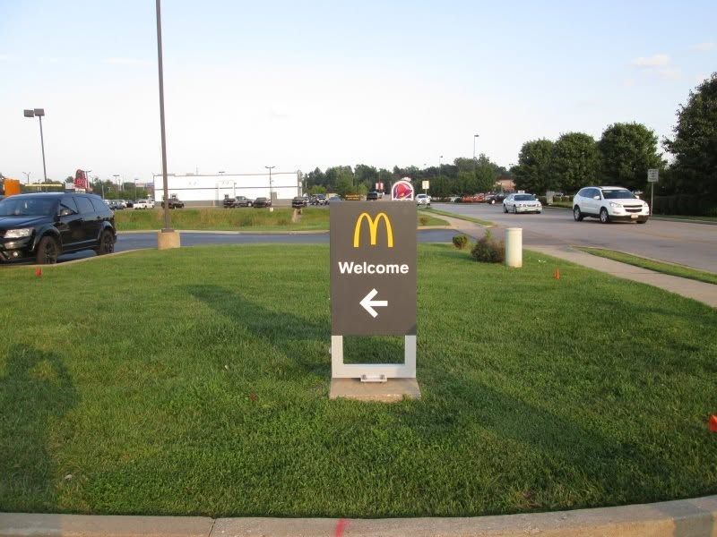 McDonald’s “Welcome” sign with left arrow on a grassy roadside median near a parking lot. McDonald’s “Welcome” sign with left arrow on a grassy roadside median near a parking lot.