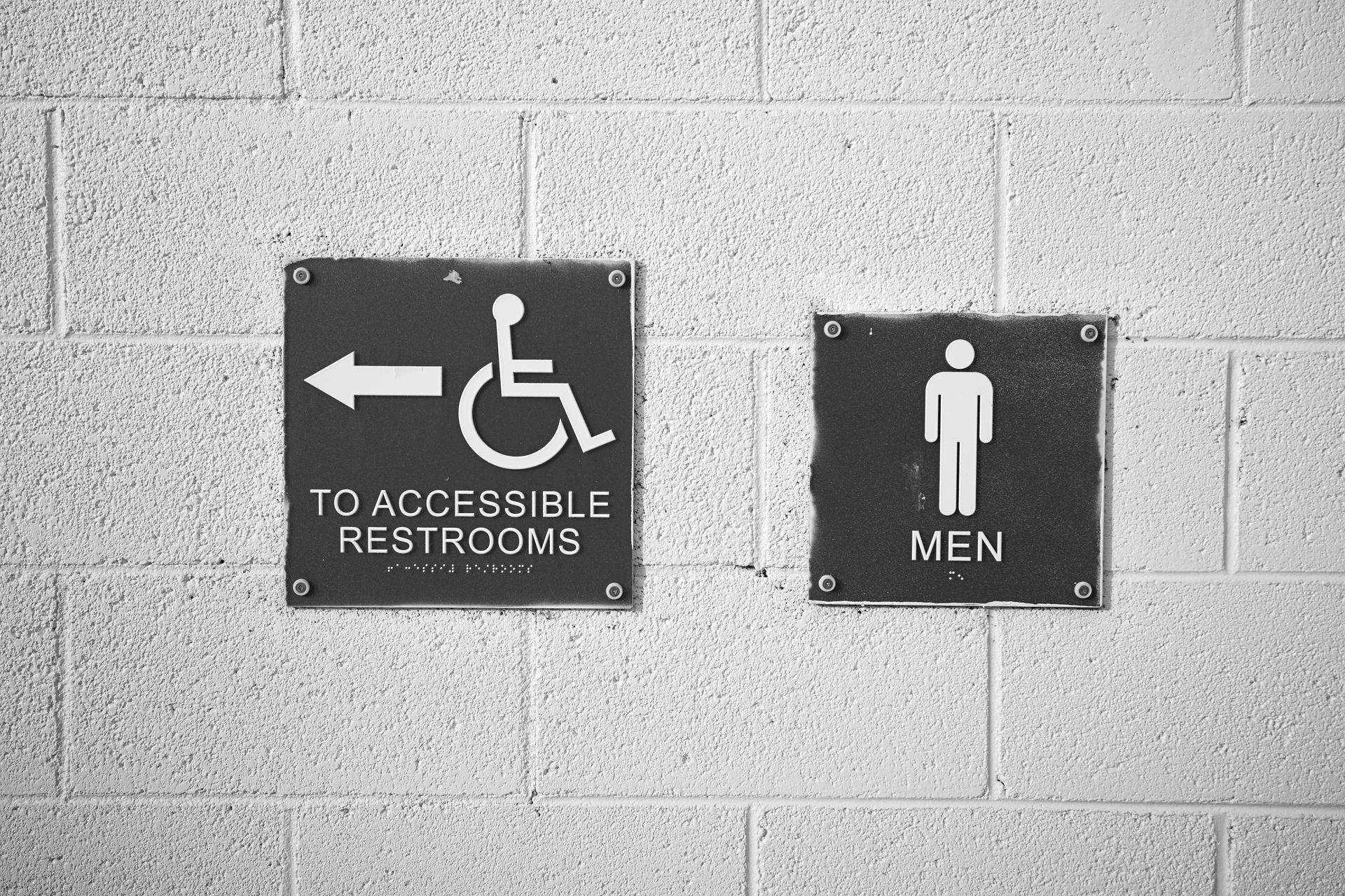 Black restroom signs on a cinderblock wall: accessible restroom with left arrow and men’s restroom icon. Black restroom signs on a cinderblock wall: accessible restroom with left arrow and men’s restroom icon.