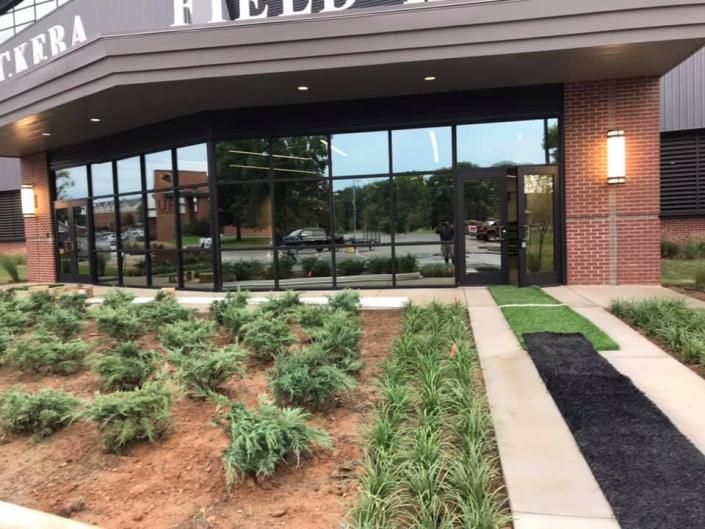 Glass-front building entrance with brick columns, landscaped shrubs, and a sidewalk path