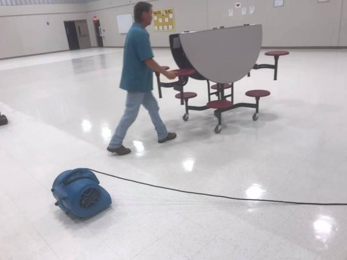 Person pushing a gray cafeteria table on a shiny floor in a large empty room; blue bag and cord in foreground
