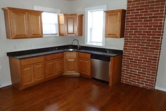 Empty kitchen with wood cabinets, black countertops, stainless dishwasher, and brick accent wall