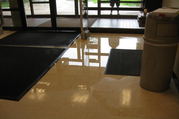 Indoor entrance with dark floor mats, reflective tile floor, and a cylindrical trash can near glass doors.