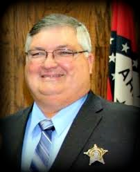 Smiling sheriff in dark suit with star badge, standing before a flag backdrop