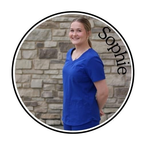 Young woman in blue scrubs smiling against a stone wall with “Sophie” text