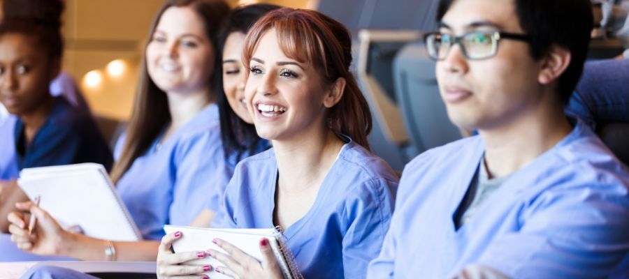 Group of nursing students in scrubs, smiling and listening attentively in a classroom setting.