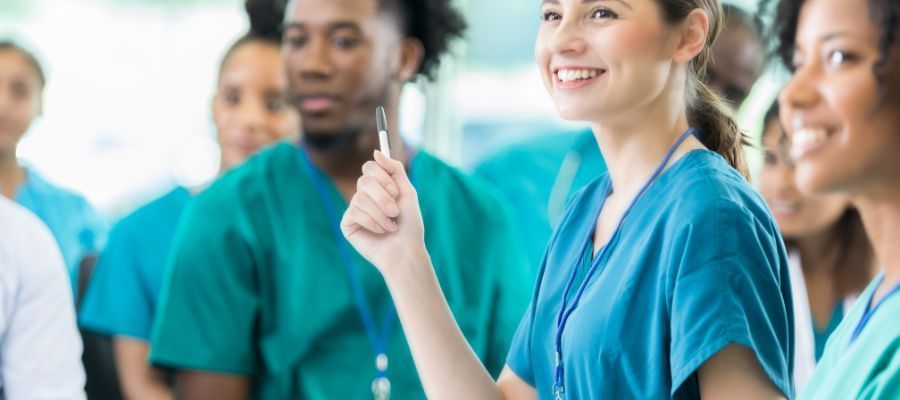 Nurses in teal scrubs smile during a meeting; a woman holds a pen up, while others look on.