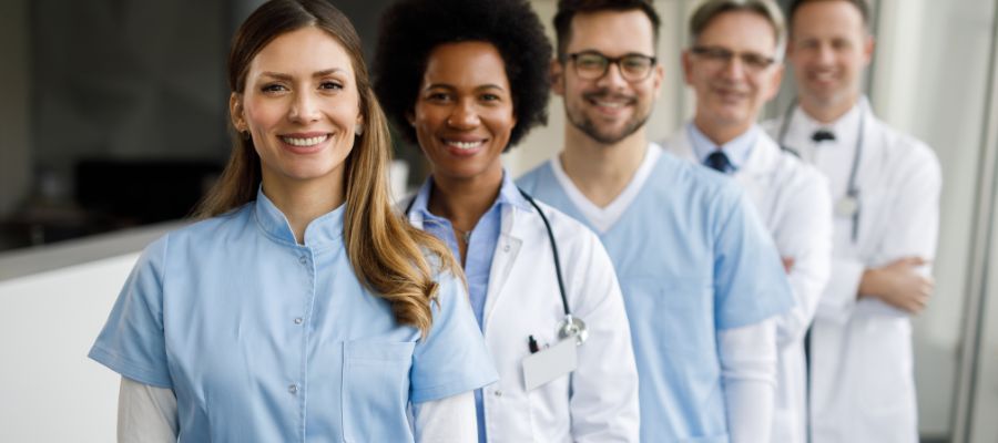 A diverse group of smiling healthcare professionals in light blue scrubs and white coats.