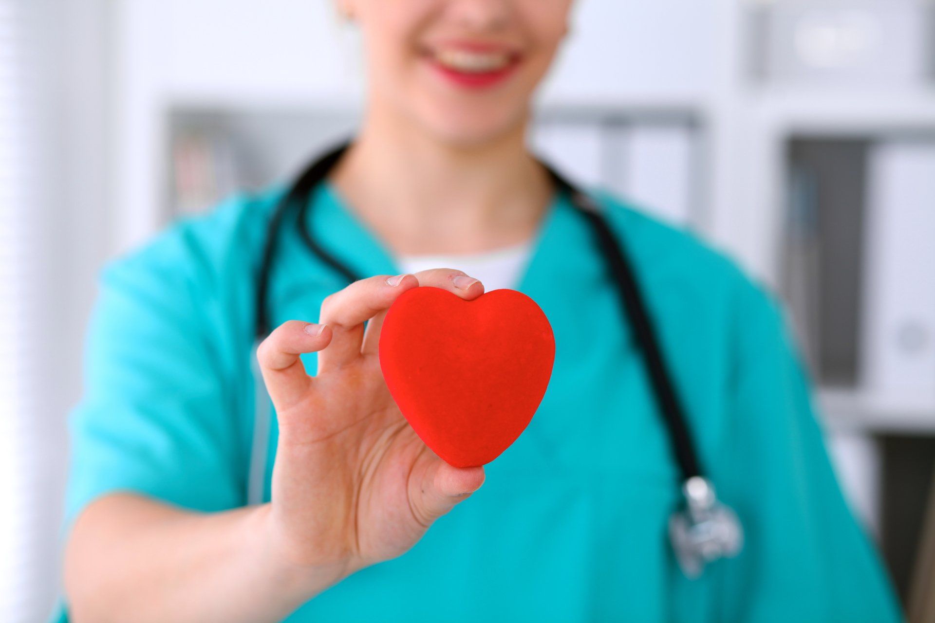 Doctor in green scrubs smiling, holding a red heart; stethoscope around neck.