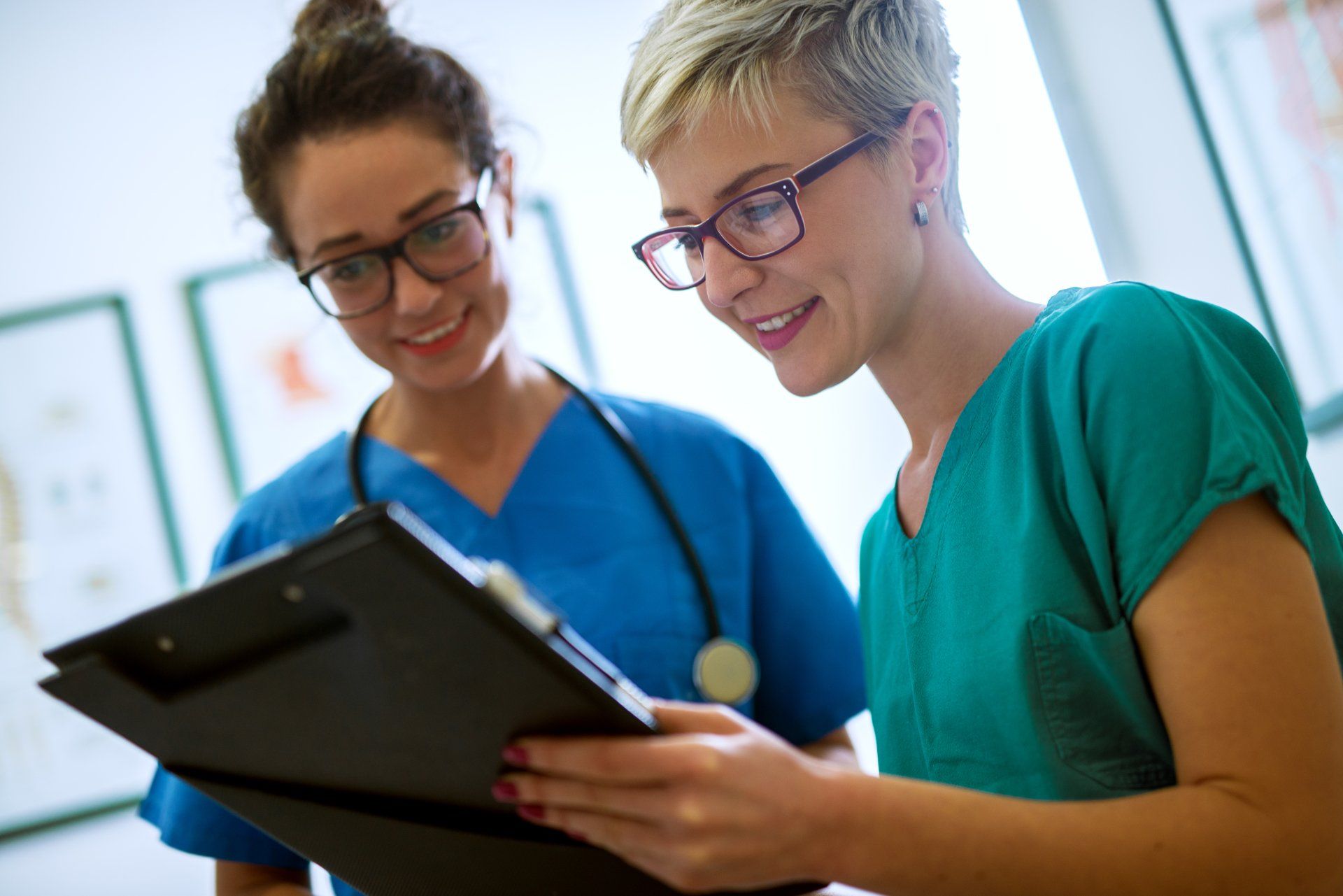 Two nurses, in blue and green scrubs, review a clipboard together, smiling.