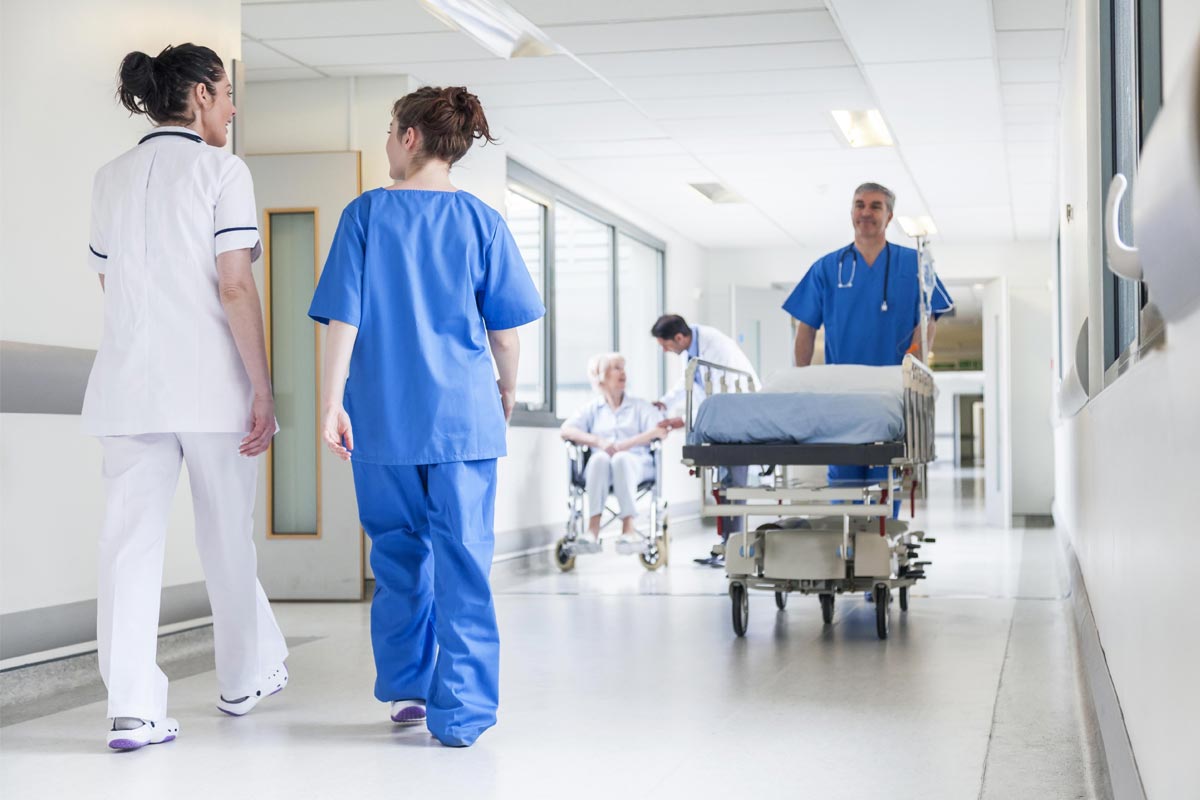 Hospital hallway with medical staff and patients; two nurses walking, a man pushing a stretcher, and a patient in a wheelchair.