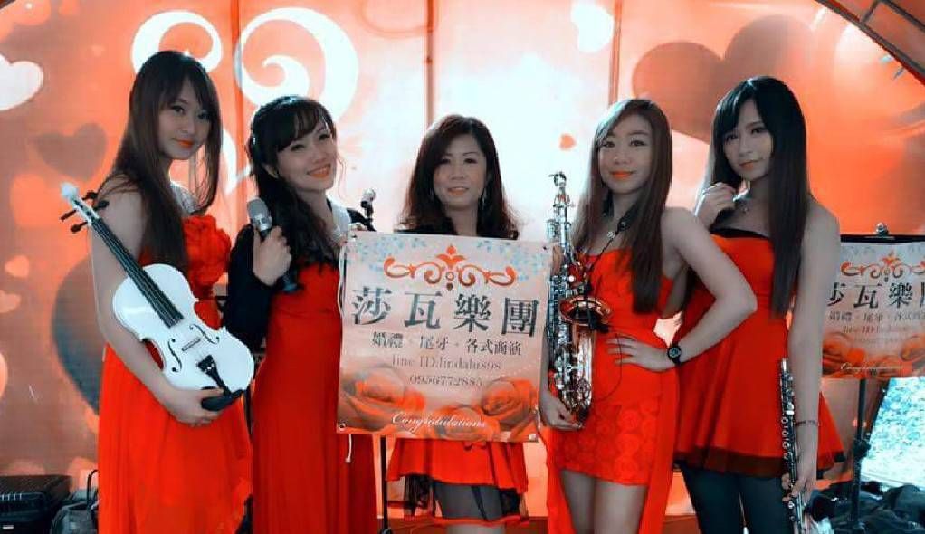 莎瓦五位身穿紅色連身裙的女子在舞台上擺姿勢，手持標語，背景是明亮的橙色。