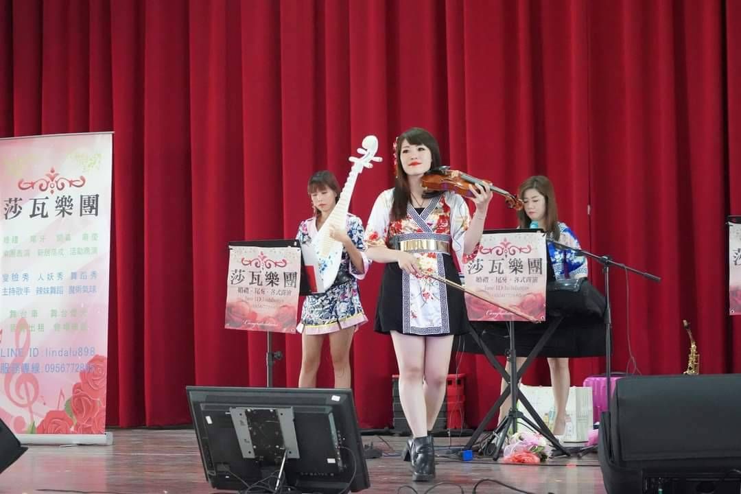 莎瓦三位表演者站在紅色布幕前的舞台上，手持麥克風和標語，眼前是一小群觀眾。