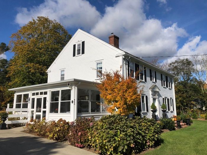 White two-story house with a porch and autumn foliage in front on a sunny day.