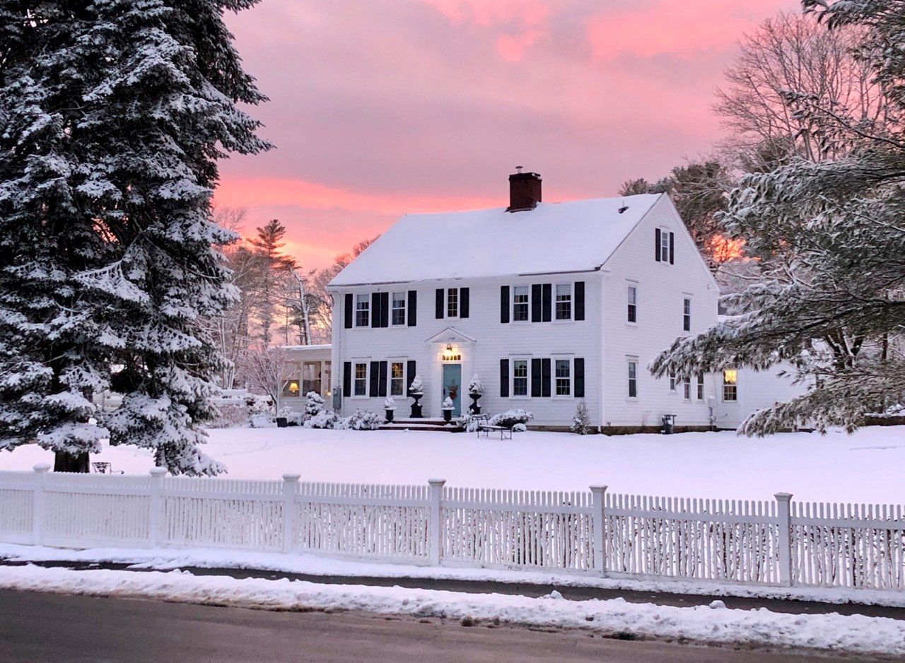 Snow-covered white house with black shutters, pink and orange sunset, white picket fence.