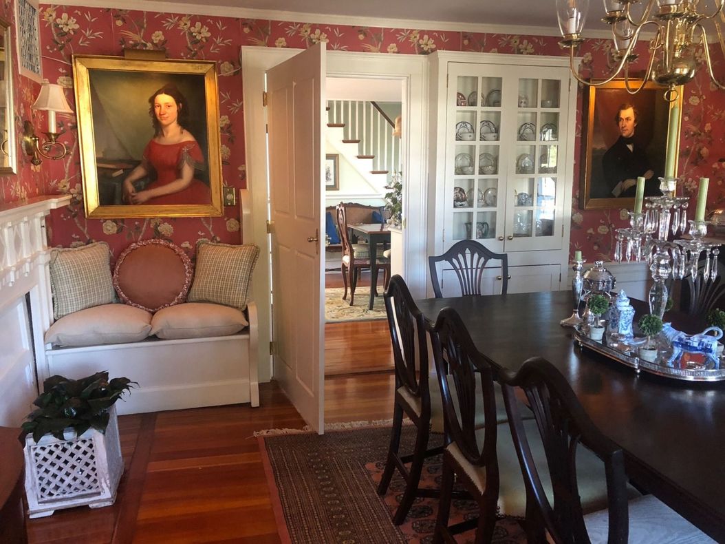 Interior with dining table, paintings, bench, and view into another room with staircase. Red wallpaper.
