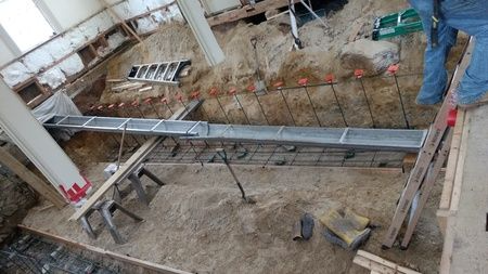 Construction site with excavated ground, rebar, metal forms, and a person in jeans.