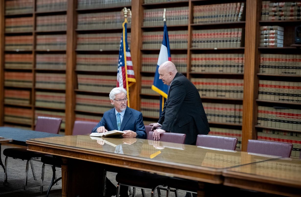 Two officials meeting at a conference table in a library with bookshelves and flags in the background