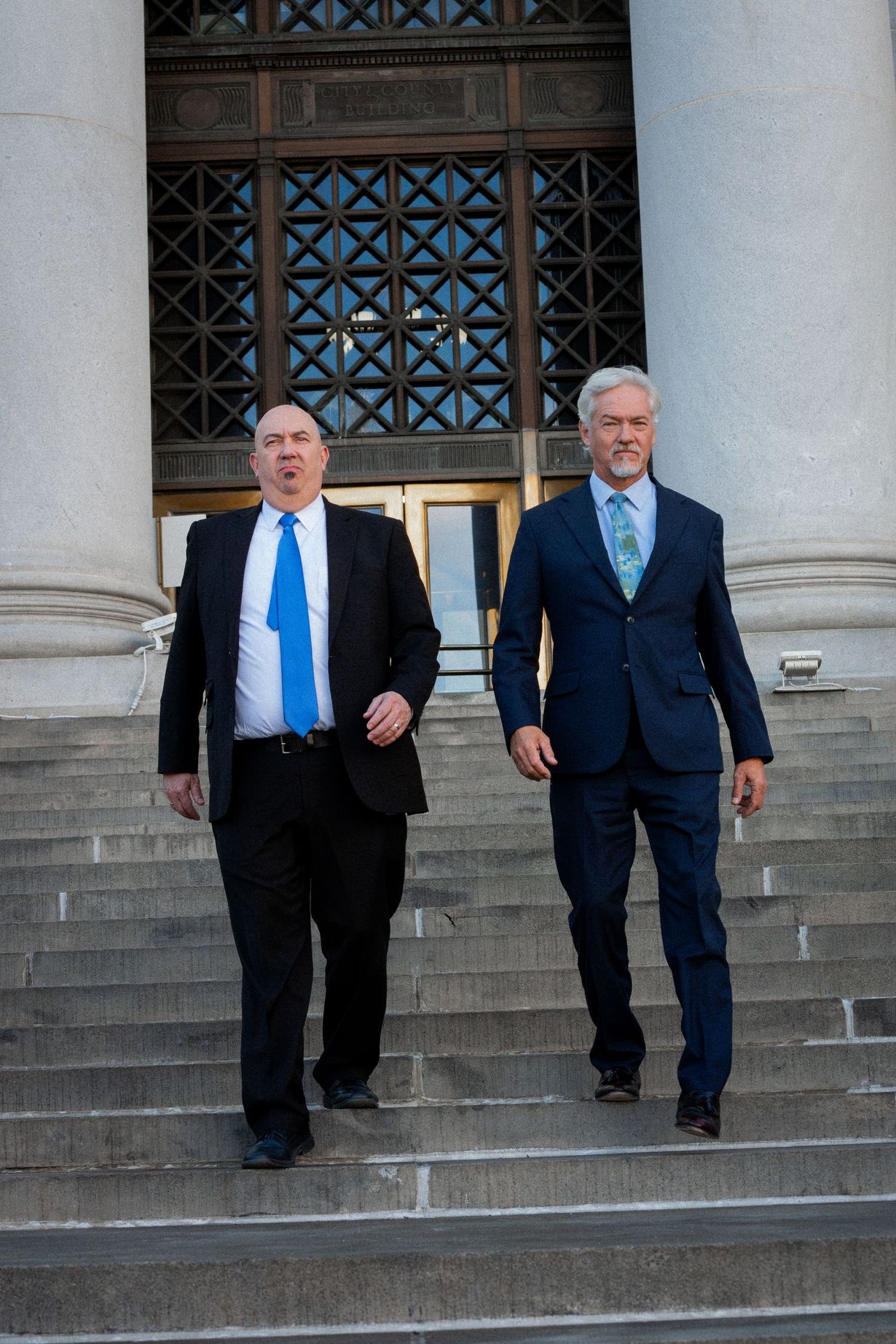 Two men in suits walking down courthouse steps, one in a blue tie and one in a dark blue tie.