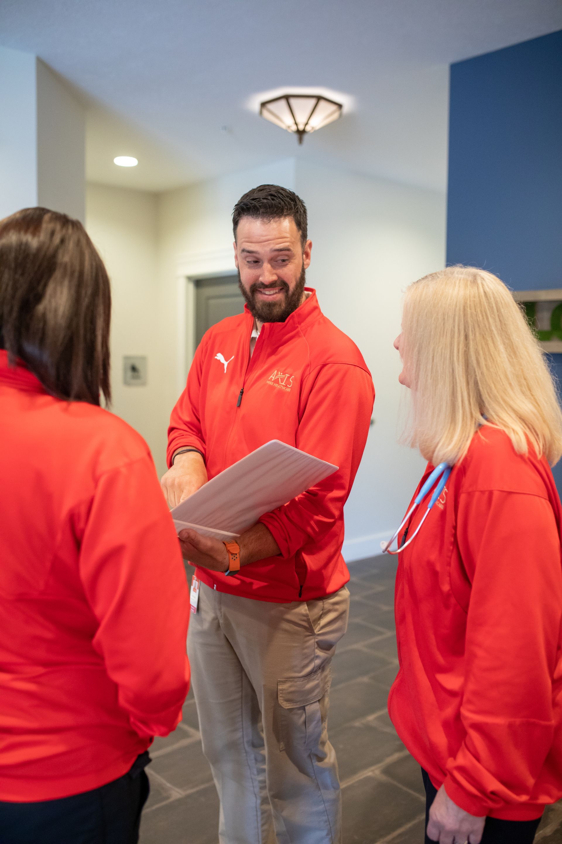 Three people wearing red jackets stand in an indoor office space, talking and looking at a document held by a man.