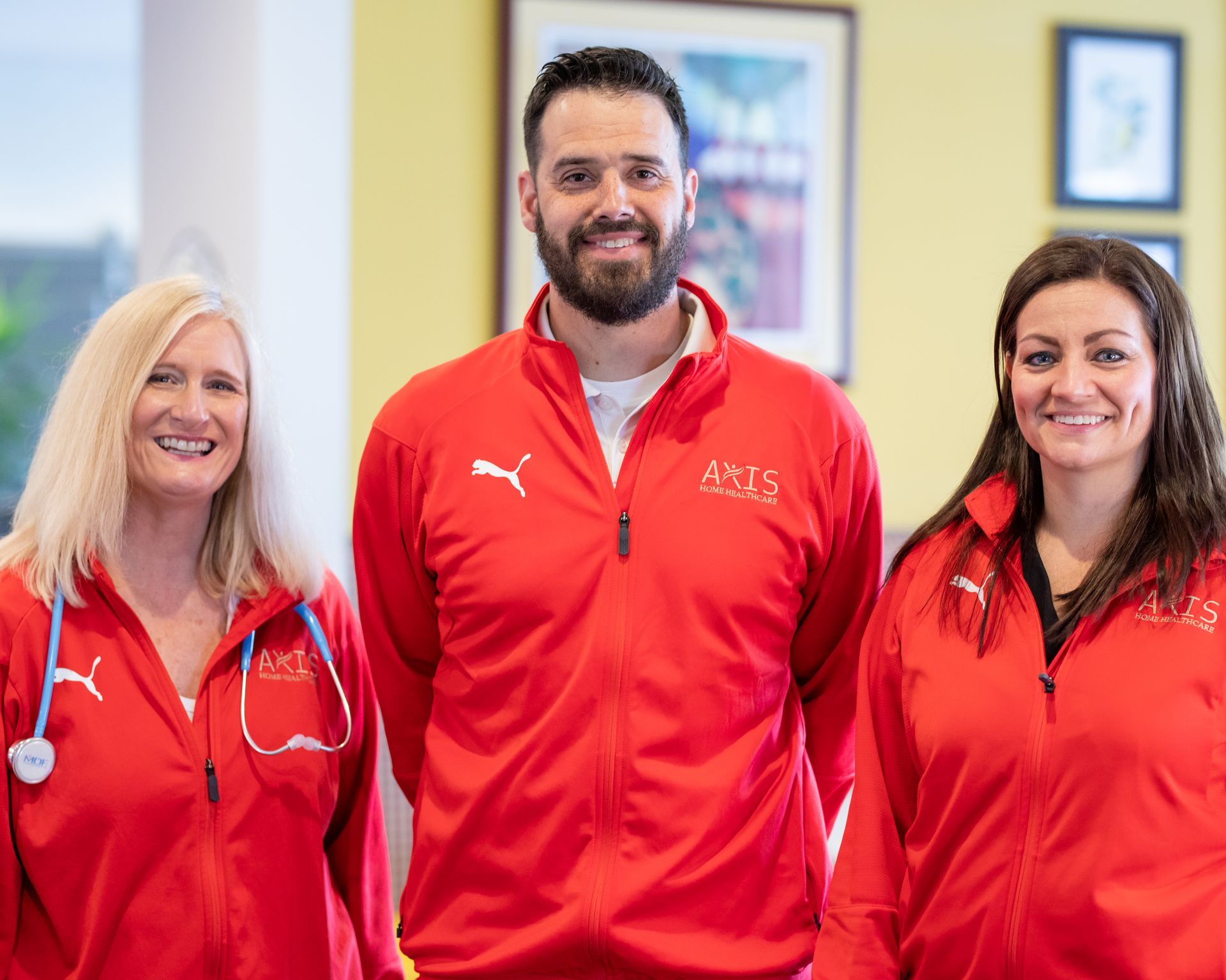 Three people wearing matching red Puma jackets with an Axis logo stand smiling in an office with framed wall art.