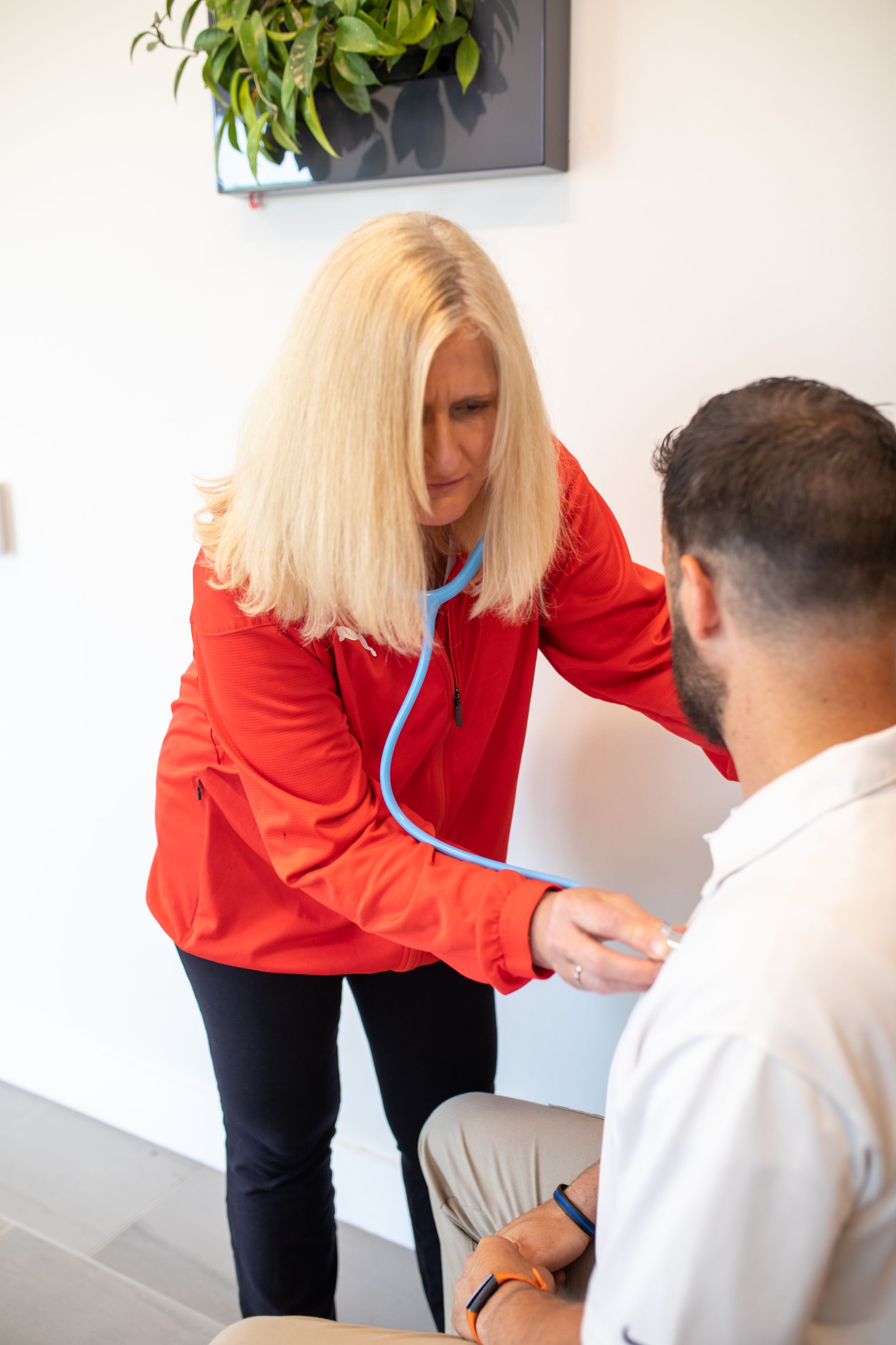 A medical professional in a red jacket uses a stethoscope to examine a patient's chest in a bright, modern clinic.
