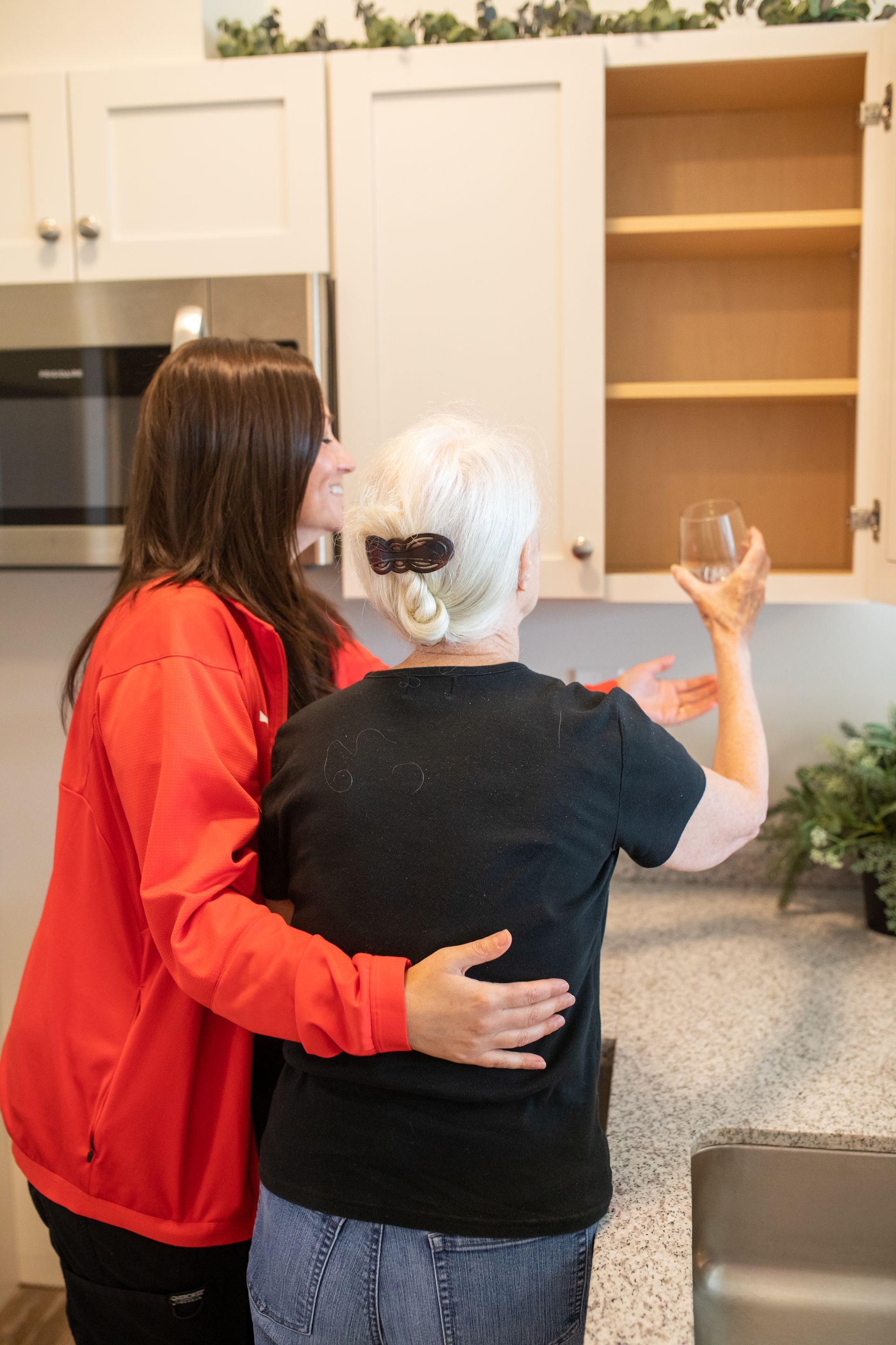 A person in a red jacket supports someone in a black shirt, who is placing a glass into an empty kitchen cabinet.