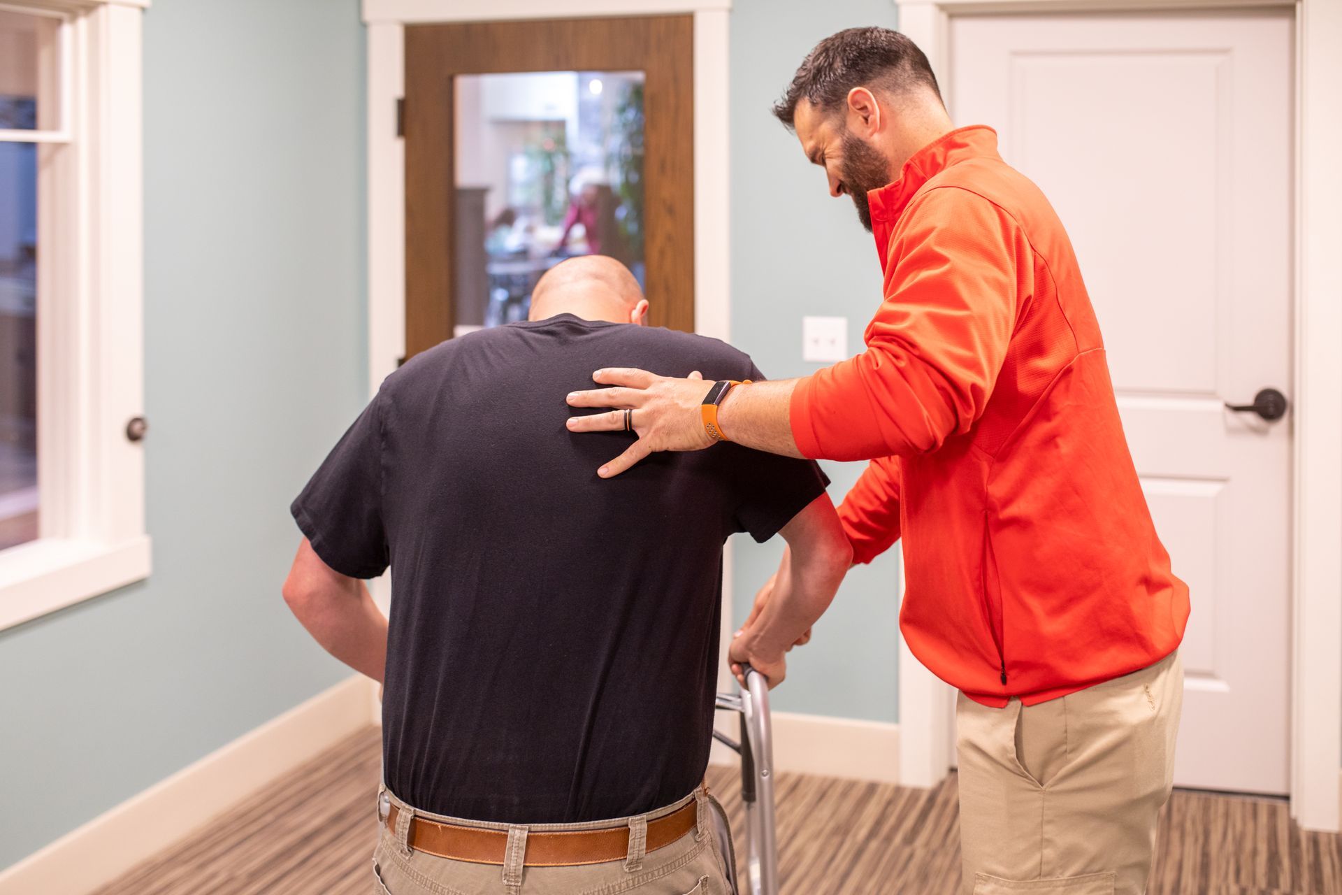 A physical therapist in an orange jacket assists a patient using a walker in a room with light blue walls.