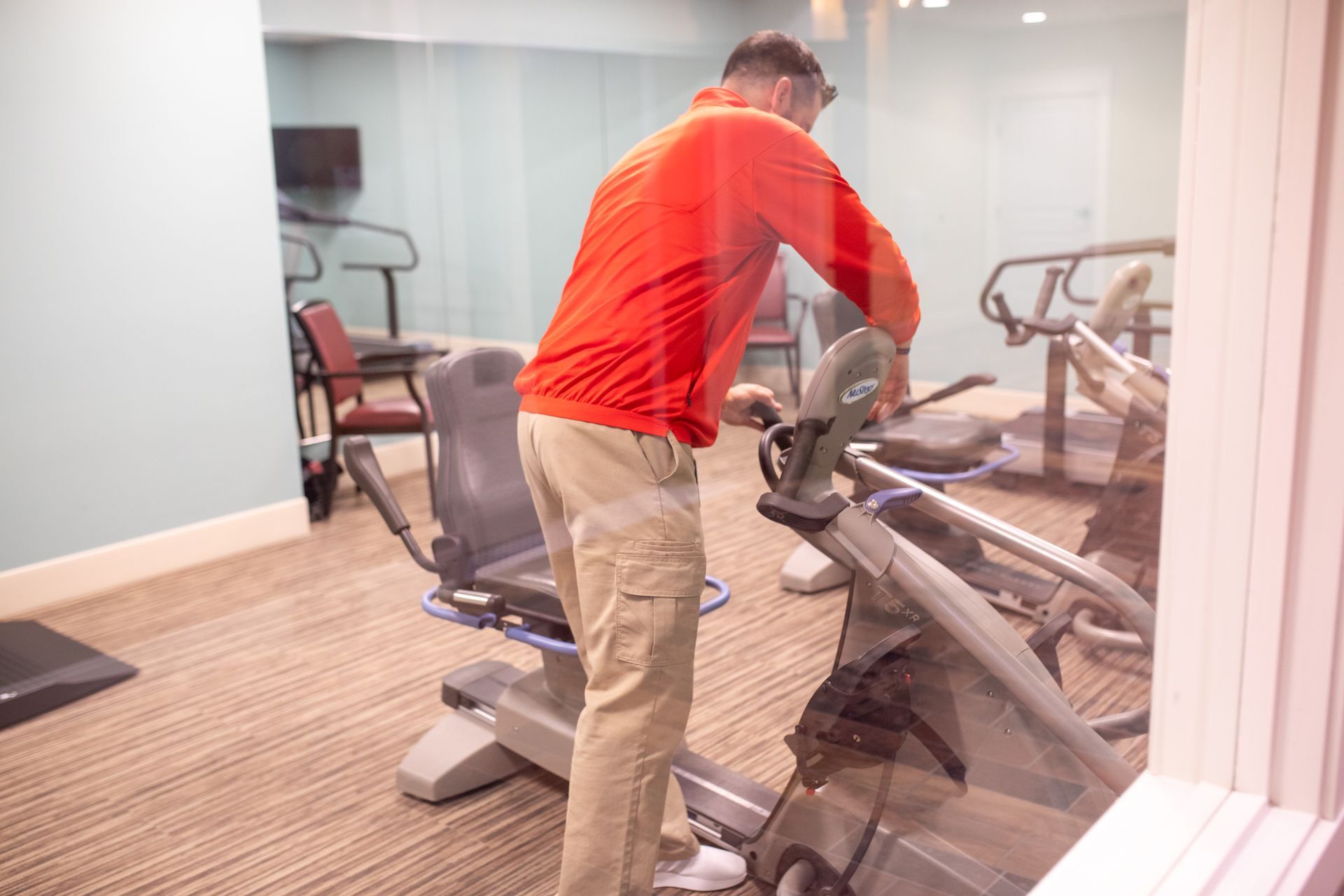 A person in a bright orange jacket and khaki pants stands in a gym, adjusting the settings on a stationary exercise bike.