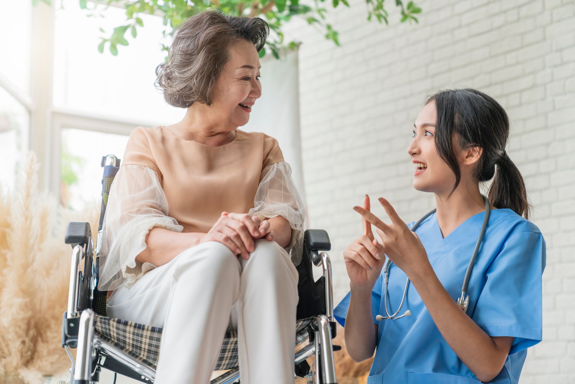 A healthcare worker in blue scrubs speaks with a person in a wheelchair, gesturing while they interact in a bright room.
