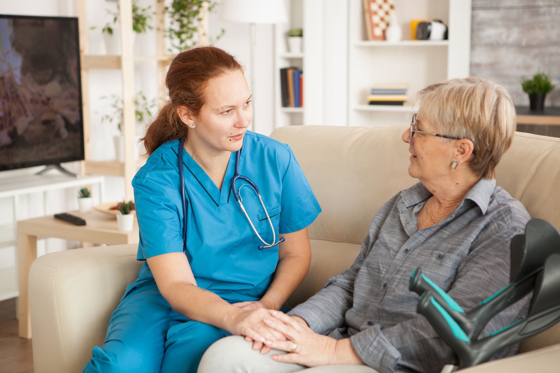 A medical professional in blue scrubs sits on a couch holding hands with an individual using forearm crutches.