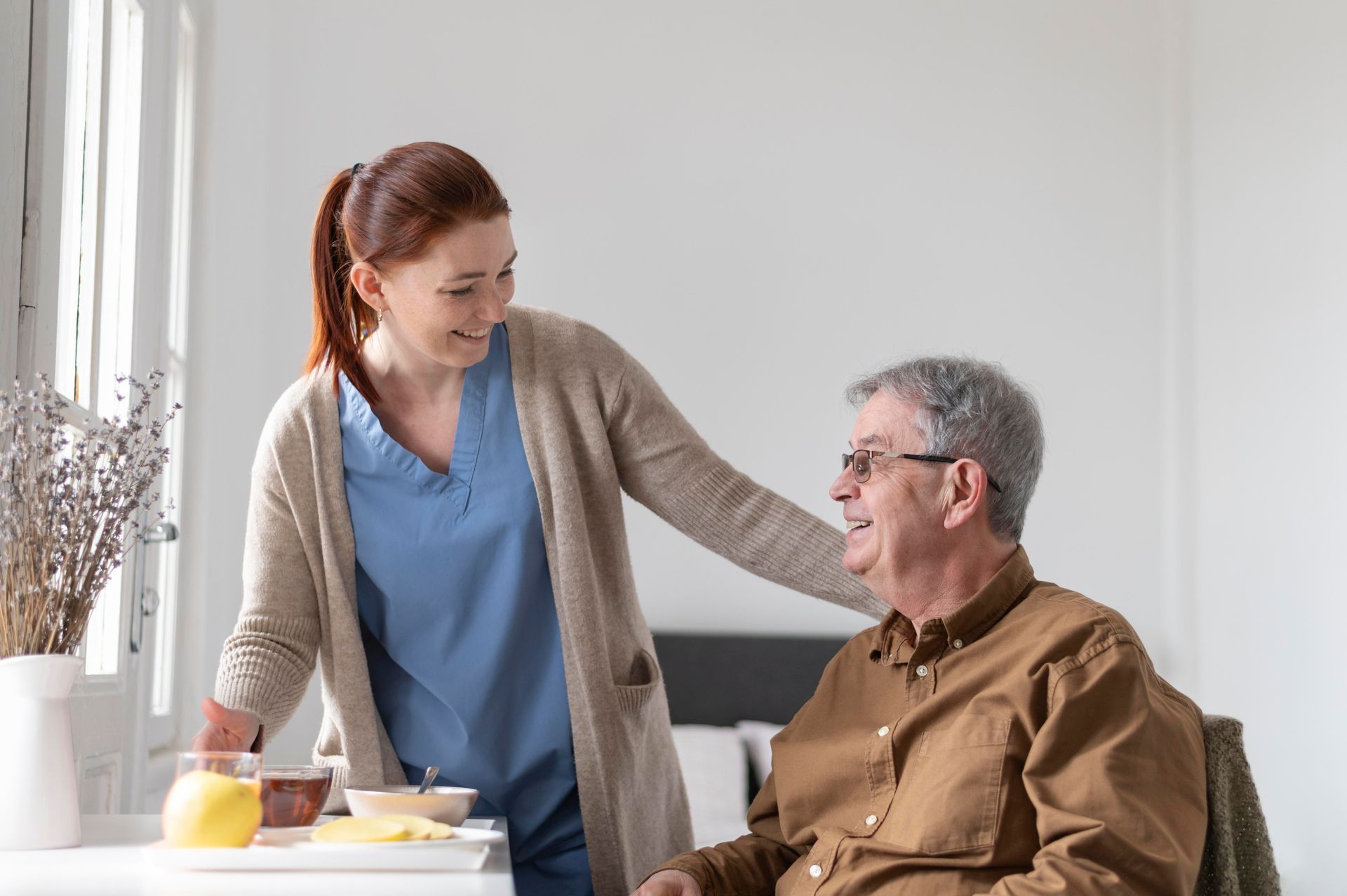A caregiver in blue scrubs and a cardigan smiles while talking to a person sitting at a table with breakfast.