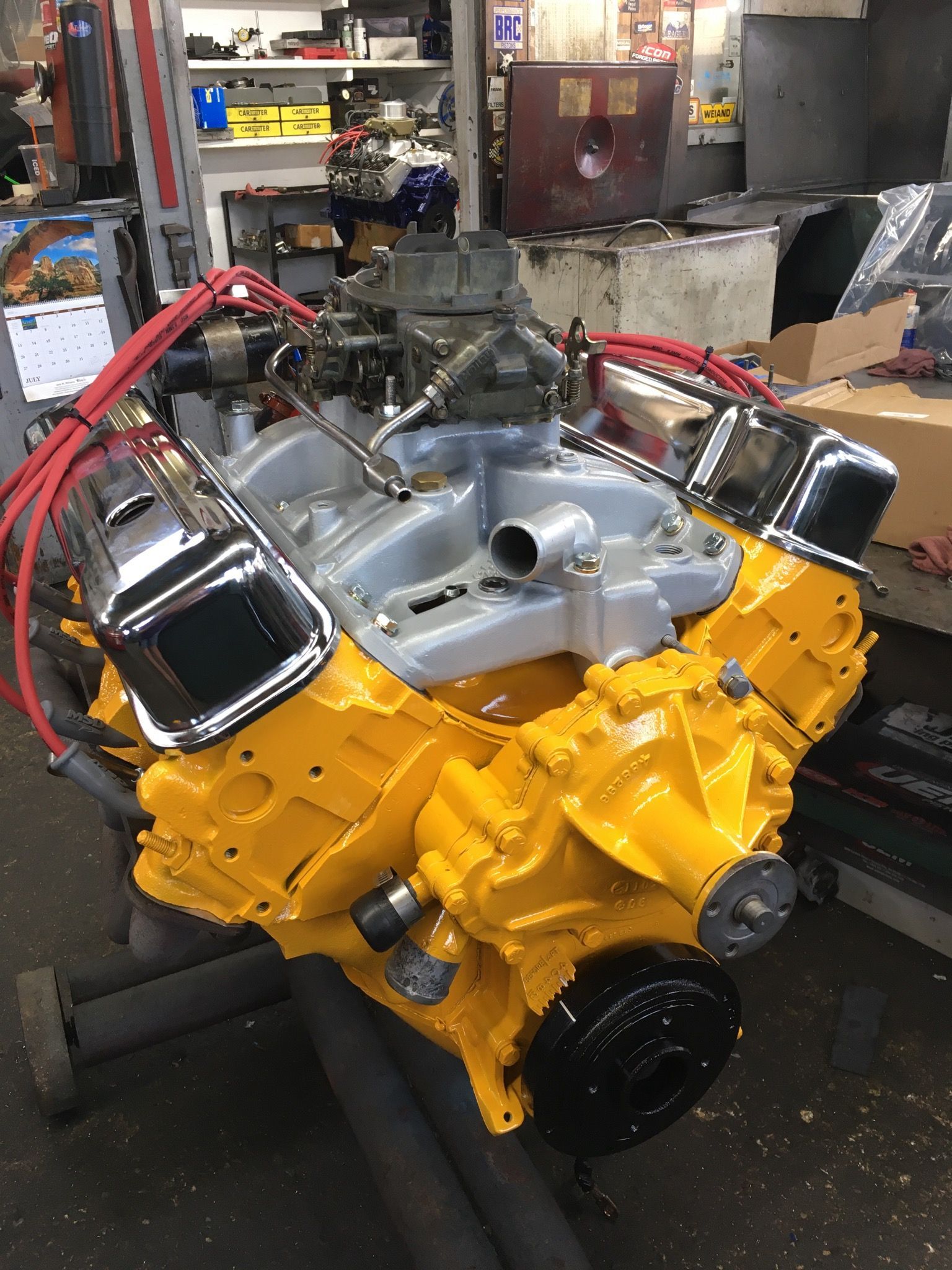 Yellow and gray painted car engine with chrome valve covers, sitting in a garage.
