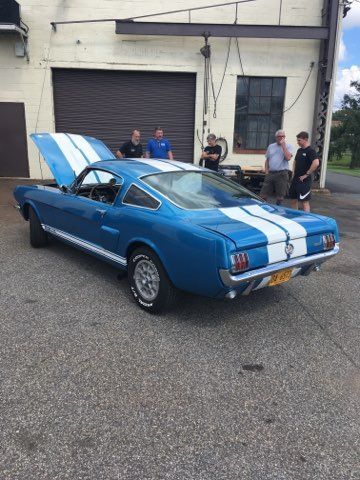 Blue classic muscle car with white racing stripes parked outside a garage, with people standing nearby