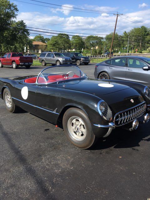 Black vintage Corvette roadster with racing roundels, parked outdoors.