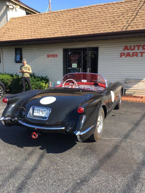 Black vintage Corvette parked outside an auto parts store with a person standing nearby.