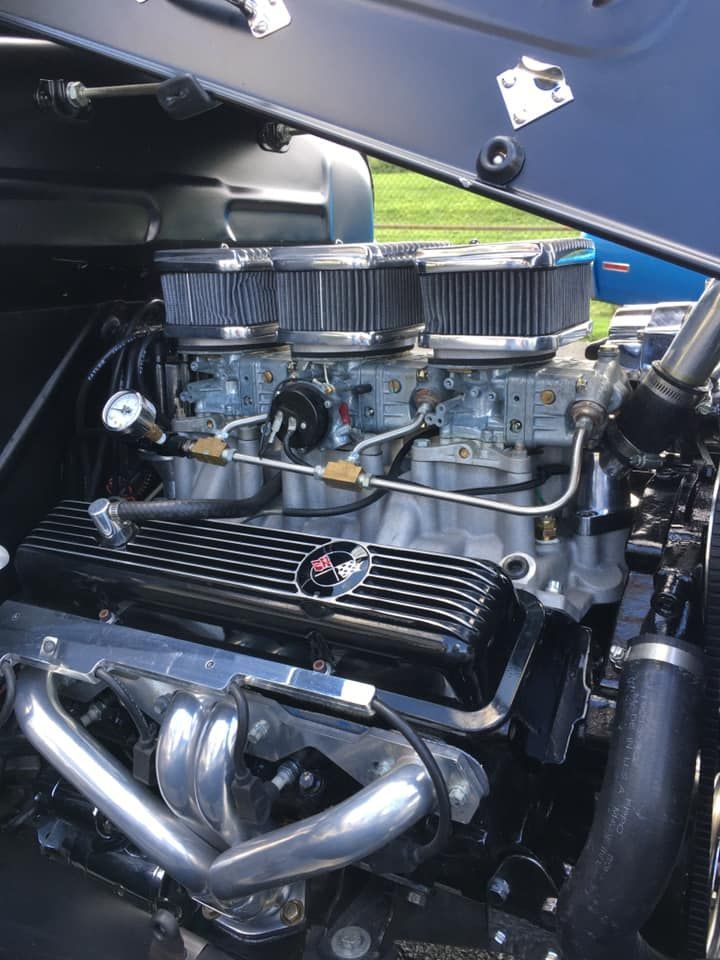 Close-up of a hot rod engine with three carburetors, chrome headers, and black valve covers.
