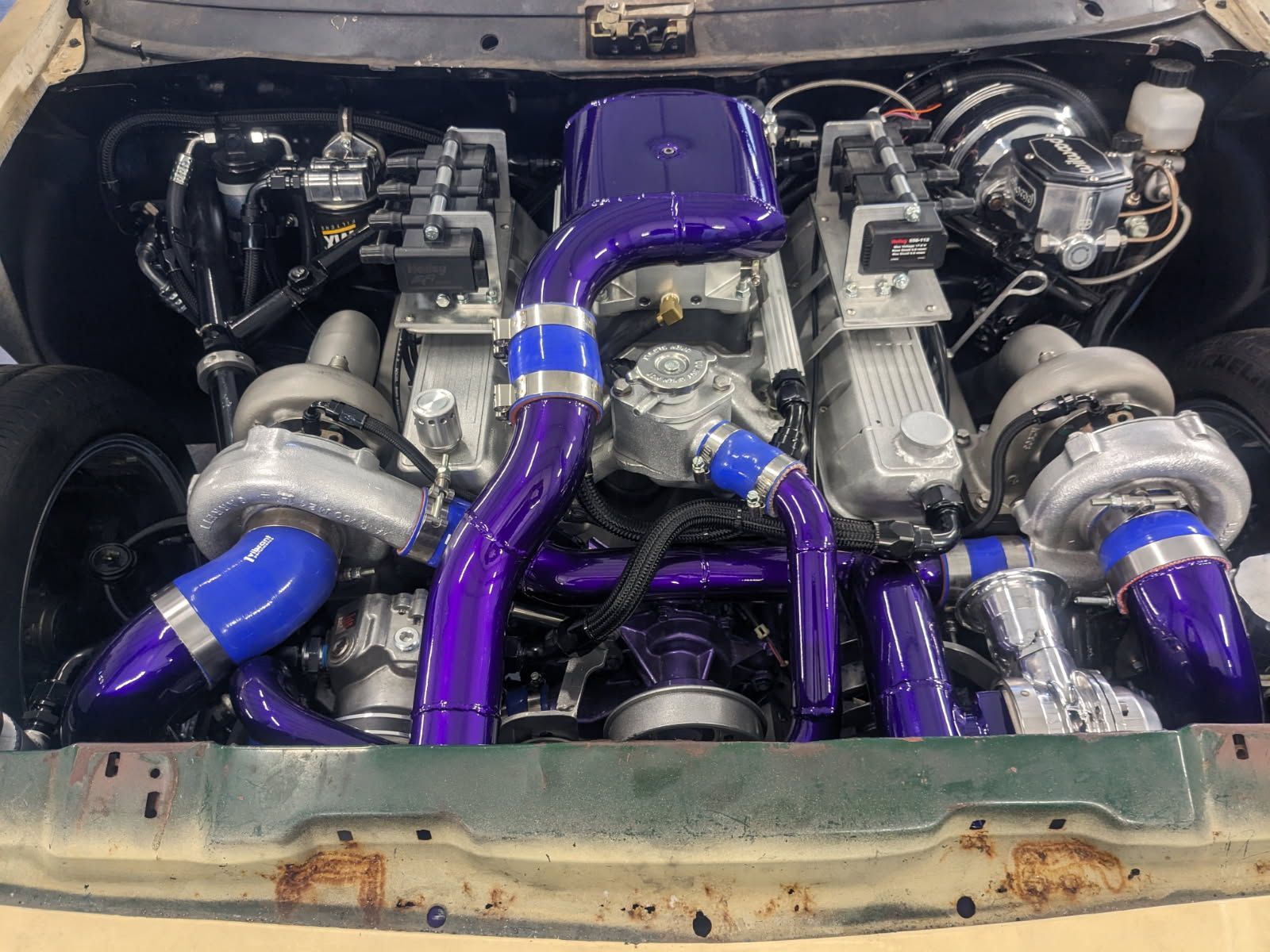 Car engine bay with twin turbos, purple piping, silver turbos, and a metallic finish.